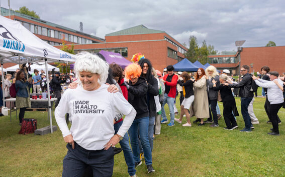 People dancing the Finnish line dance "letkajenkka" outdoors with university buildings in the background.
