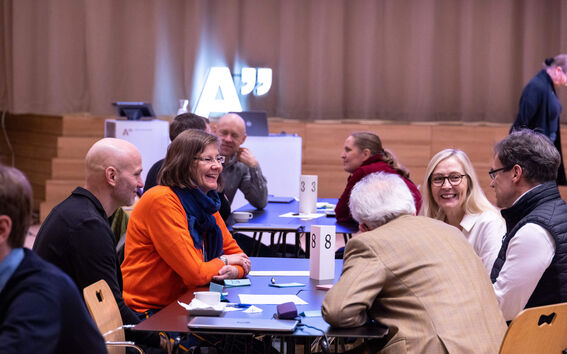 Ihmiset istuvat kokoushuoneessa numeroiduilla pöydillä. Taustalla näkyy kirkas 'A'-symboli.
