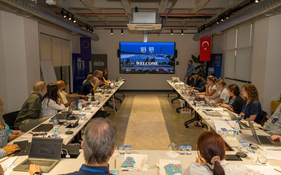 A conference room with attendees seated around a U-shaped table, facing a screen displaying 'SUN' and 'WELCOME'.