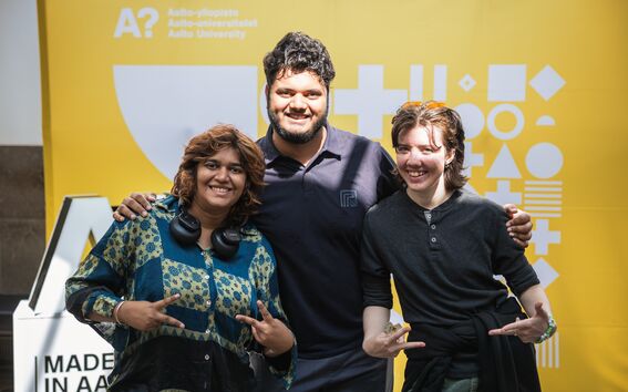 Three people with arms around each other in front of a yellow Aalto University backdrop. One is wearing headphones.