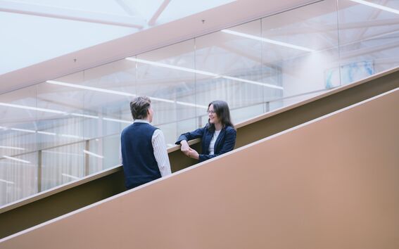 Kaksi ihmistä keskustelemassa modernilla portaikolla, taustalla lasiseinä ja kattovaloja.
