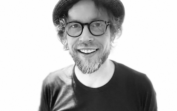 A man wearing a black T-shirt and a hat, with an arm tattoo, shown against a plain white background.