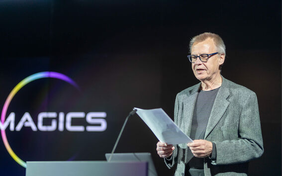 Vanhempi mies harmaassa bleiserissä pitää papereita, seisoo podiumin lähellä, taustalla sana 'MAGICS'.