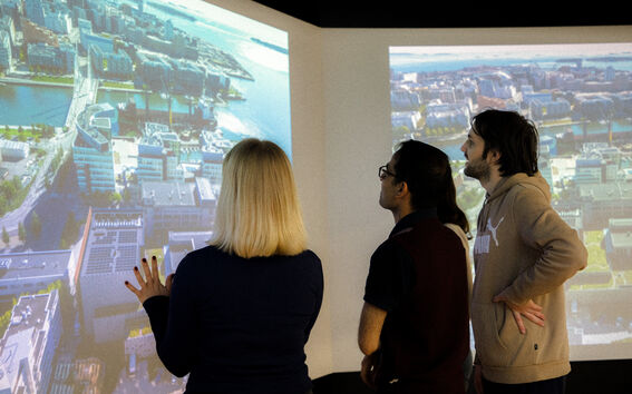 Kolme ihmistä seisoo suuren näytön edessä, jossa näkyy kaupungin ilmakuva modernista rakennuksista ja vesiväylistä.