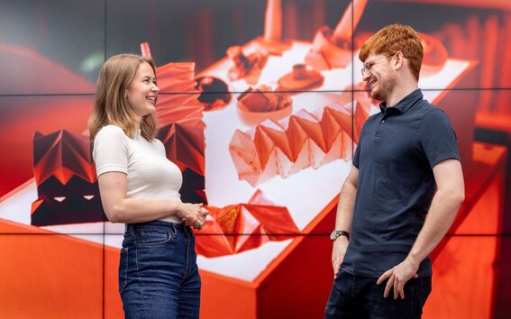 Two people standing in front of a large screen displaying geometric, red and white shapes.