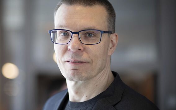 A person wearing a grey suit and black shirt. The background is blurry and appears to be indoors.