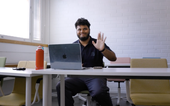 Person with laptop on white desk in classroom. Colourful chairs, red bottle, large window.