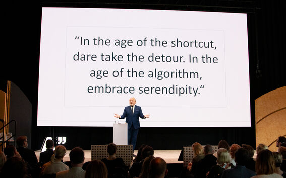Speaker in a suit gesturing on stage, large screen on the background