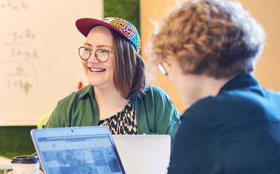 Kaksi henkilöä keskustelemassa pöydän ääressä, jossa on kannettavia tietokoneita ja kahvimuki ja taustalla on taulu.