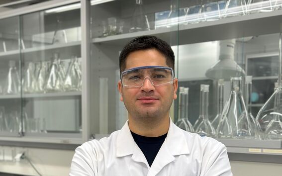A person in a white lab coat stands in front of laboratory shelves filled with glass containers.