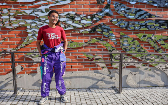Hitesh Monga wearing Tutor in Aalto University shirt and overalls, standing in front of a brick wall with metal artwork