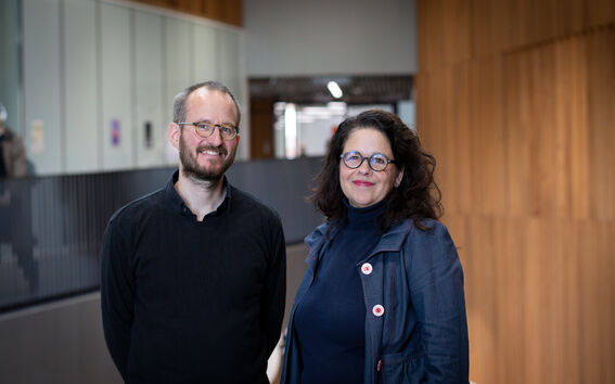 Kaksi ihmistä seisoo sisällä kaiteen vieressä. Miehellä musta paita, naisella tummansininen takki.