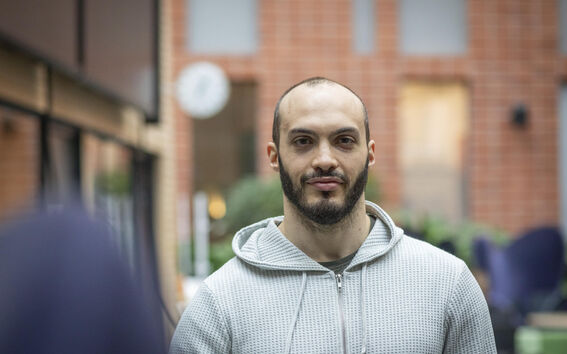 A person wearing a light grey hoodie stands indoors with a brick wall and green plants in the background.