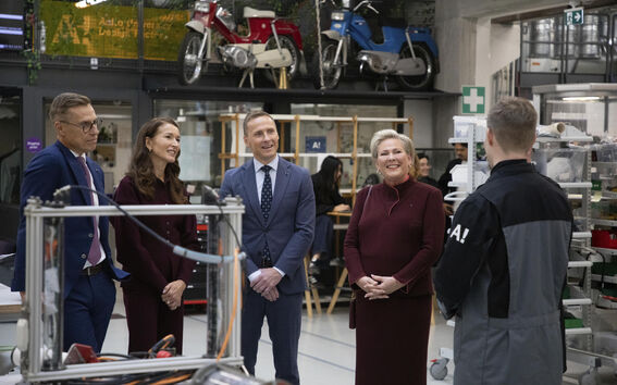A group of people in formal clothing are visiting a modern workshop. Vintage motorbikes are displayed on a high shelf.