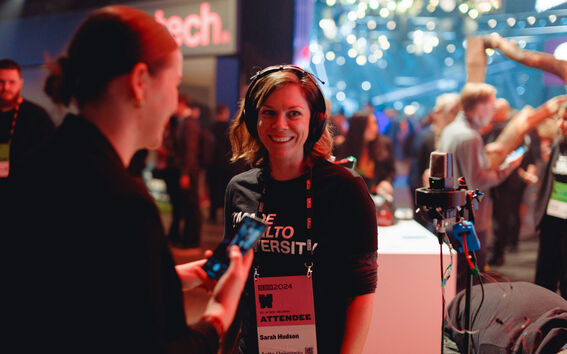 People at a tech event, one wearing an attendee badge and headphones. Sign reads 'tech' in background.