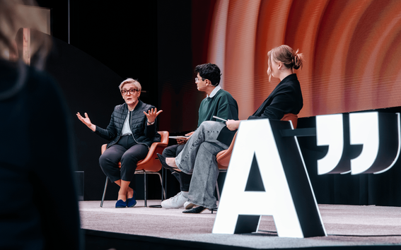 Kolme ihmistä istuu tuoleilla lavalla, jossa on suuri valaistu kirjain 'A' ja lainausmerkit heidän vieressään.