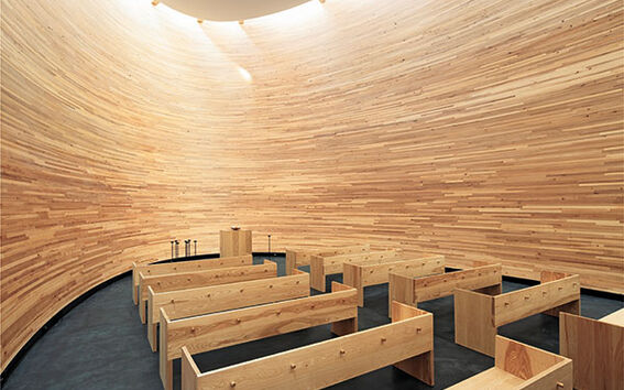 Modern chapel with curved wooden walls, wooden benches, and an altar under a round, bright ceiling.