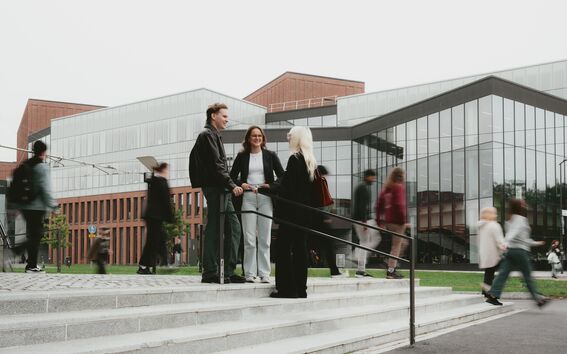 Three individuals conversing on steps in front of modern glass buildings, with pedestrians walking by.