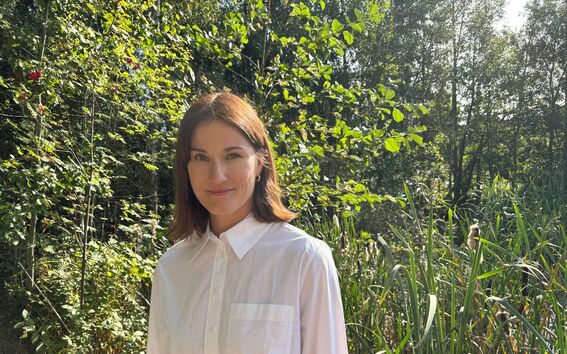 Iiris Hörhammer seisoo ulkona valkoinen paita päällään. Taustalla korkeaa ruohoa ja puita.