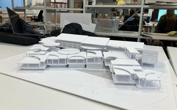 Scale model of a modern building with geometric shapes on a table in a classroom, students working in the background.