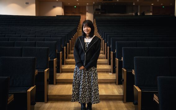 A person standing in an empty auditorium wearing a floral dress and black jacket with wooden seats.