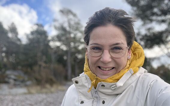 A person wearing a white coat and yellow hoodie stands outside in windy weather, with forest in the background.