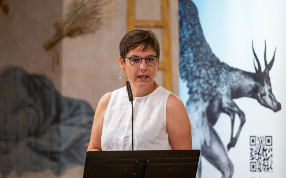 Person in a white blouse speaks into a microphone, presentation slides projected on the wall behind.