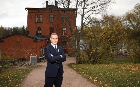 Person in a dark suit stands on a gravel path in front of an old brick building surrounded by autumn trees.