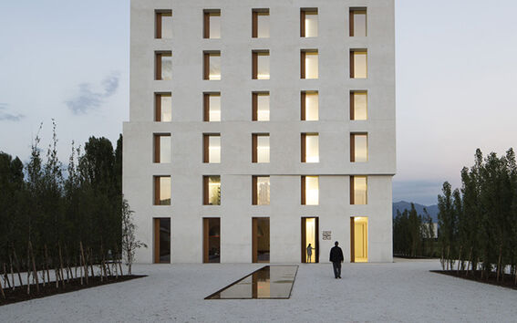 A modern, rectangular building with symmetrical windows. A lone person walks towards the entrance. Trees line the pathway.