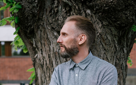 Person with crossed arms leaning on a large tree trunk, wearing a grey buttoned shirt and black trousers.