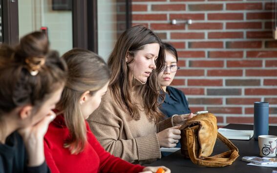 People sitting at a table with mugs, notebooks, and a brown bag. One person is peeling an orange.