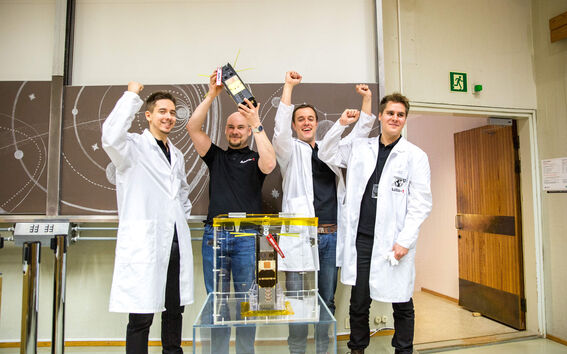 Four scientists, three in white lab coats, celebrate holding a small satellite in a laboratory.