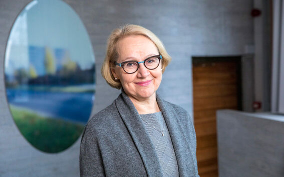 Henkilö, jolla on vaaleat hiukset, harmaa takki ja kuvioitu mekko, seisoo sisällä lähellä puuovea ja pyöreää maalausta.