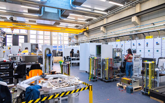 Suuri insinöörilaboratorio, jossa ihmiset työskentelevät erilaisten laitteiden parissa. Yläpuolella on keltainen nosturi ja johdot kaikkialla.