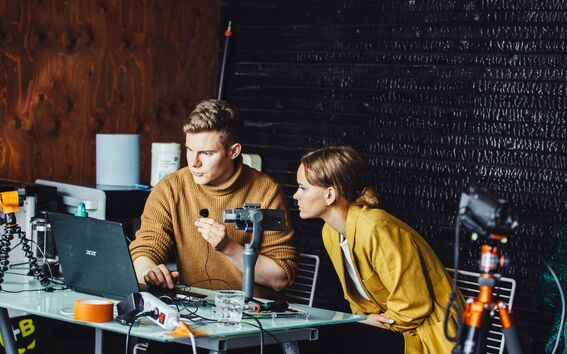 Two people working with microphones and a laptop at a table, surrounded by various electronic devices and equipment.