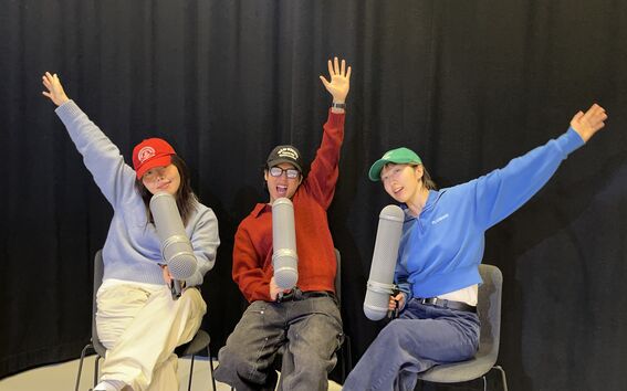 Three people sitting on chairs with large microphones, raising one arm each in a cheerful pose.