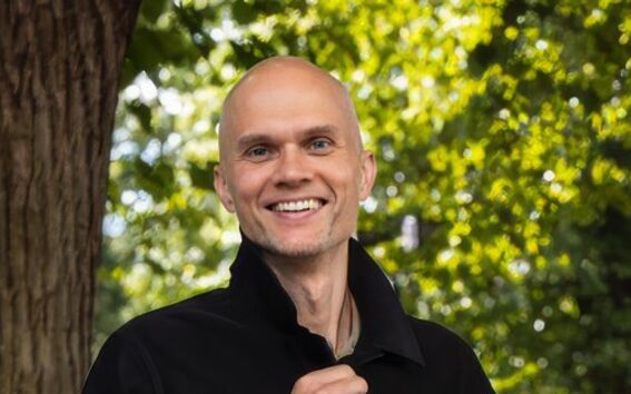 A person wearing a black jacket stands outdoors near a tree with green foliage in the background.