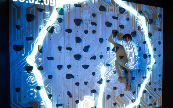 Person climbing a lit-up interactive rock climbing wall with a futuristic circuit board design and timer display.