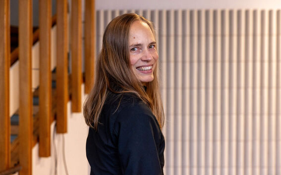 Pitkähiuksinen henkilö, jolla on tumma takki, seisoo sisällä lähellä puisia portaita ja uritettua seinää.