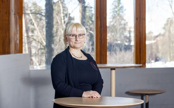 Vaaleahiuksinen henkilö mustassa asussa seisoo pyöreän puupöydän takana huoneessa, jossa on suuret ikkunat.