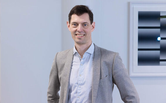 A person in a light blue shirt and grey blazer stands with one hand on their hip. A modern artwork is visible in the background.