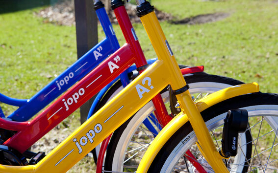 Three colourful bicycles: yellow, red, and blue, parked on grass. Each has 'jopo' written on the frame.
