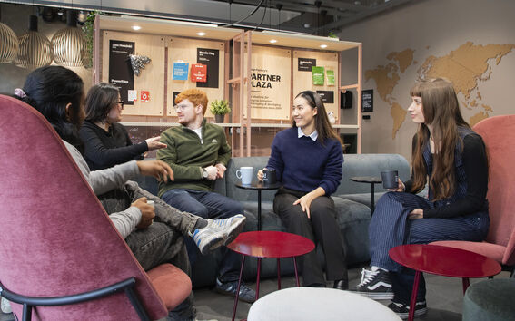 A group of people sitting in a modern lounge area with a world map on the wall and shelves displaying various items.