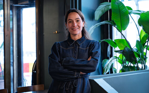 Henkilö tummassa farkkutakissa seisoo kädet ristissä huoneessa, jossa on suuret ikkunat ja vihreitä kasveja.