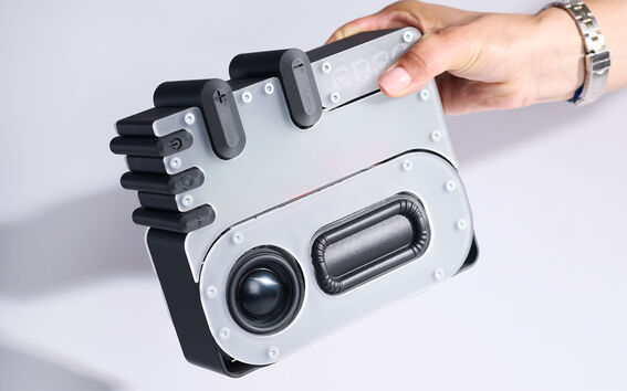 A hand holding a silver and black electronic device with buttons and a speaker on a light background.