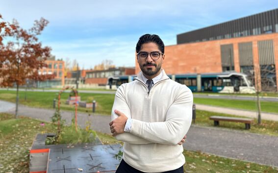 A person stands with crossed arms in front of a modern building and a bus. Trees with autumn leaves are in the background.