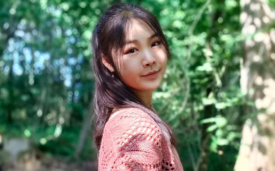 Pitkähiuksinen henkilö, jolla on vaaleanpunainen neulepaita, seisoo metsässä vihreän kasvillisuuden keskellä.