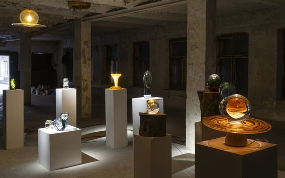 Art installation in a dimly lit room with various glass sculptures on pedestals and wooden bases.