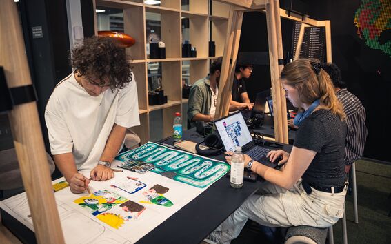 People working at a table with laptops and drawings. One person is sketching, another is typing on a laptop.