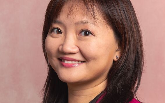 A person with dark hair wearing a magenta blazer and a black top, with a necklace. Background is pink.
